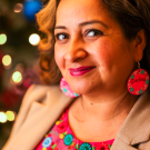 Dr. Lina Mendez wears a red top with embroidered flowers on the top and a khaki blazer. She wears two large decorative earrings.