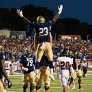 College football players celebrate; player #23 leaps with arms raised in packed stadium