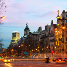 Photo of Barcelona street at dusk with the sun setting and cars driving by. A blue banner on the left reads "Tapas Night in Barcelona, Hosted by the UC Trust, Thursday, February 12th, 19:00 (Spain), Brunaqui Caffee Cucina, Barcelona"