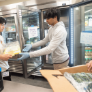 Three students in front of a fridge, the student in the middle handing food to the student on the left, and the student on the right holding a box with produce in it