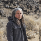 A person with long, wavy hair stands in a rocky, desert-like landscape.