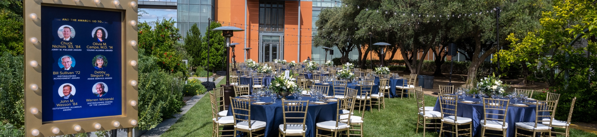 Round tables with blue linens and tablecloths, as well as gold chiavari chairs set up at the Good Life Garden for the Alumni Awards.