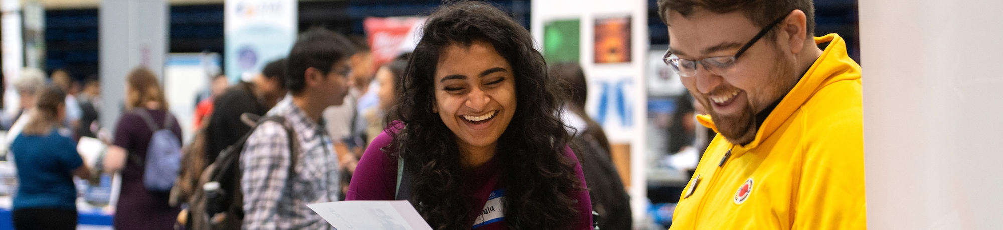 Two people smiling while laughing when looking a paper at a career fair with visible booths behind.