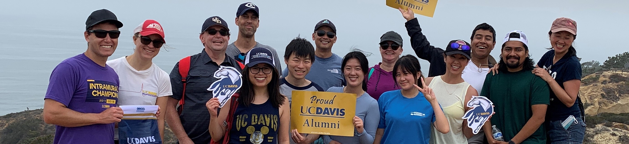 Group of UC Davis alumni holding up "Proud alumni" signs and wearing Aggie merch at the beach.