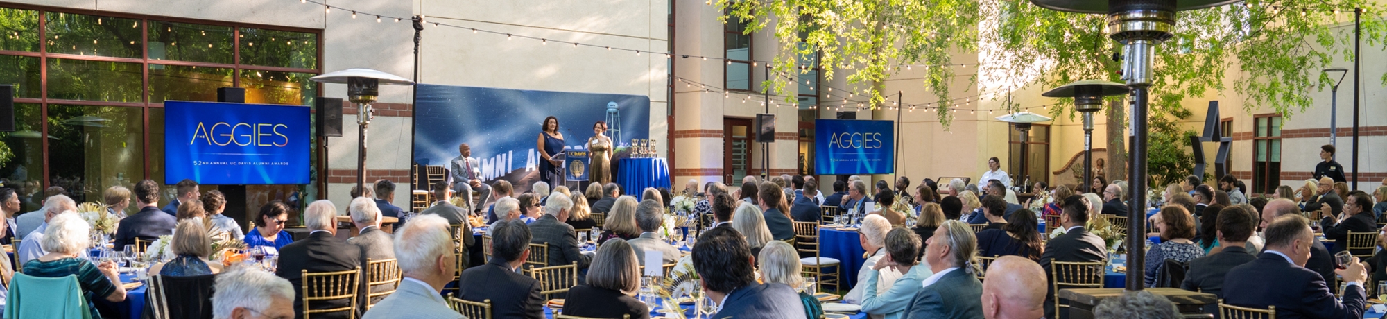 Attendees seated at a formal awards gala on gold Chiavari chairs with blue linens on round tables. The stage is visible with Chancellor Gary May seated while AVC Dana Allen and CAAA President Anu Johl Singh present at the podium.