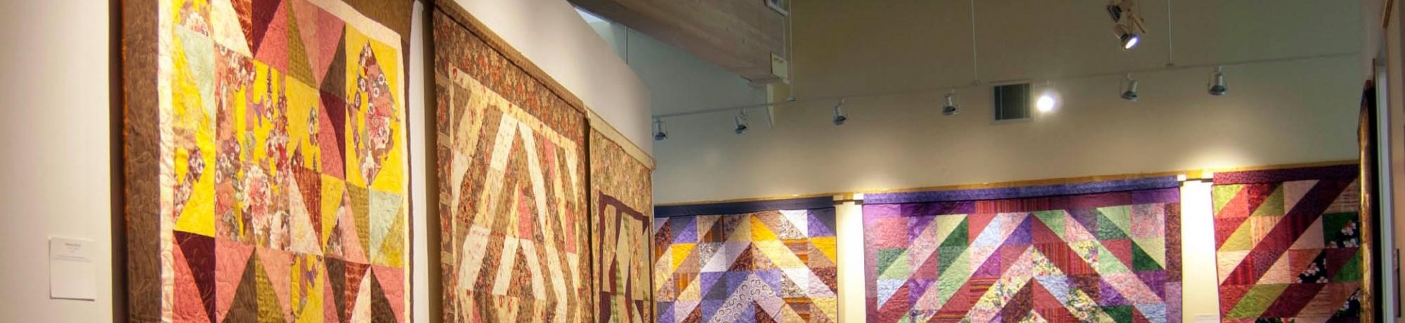 Quilts of different colors and patterns hanging on the walls of the art gallery at the Alumni Center.